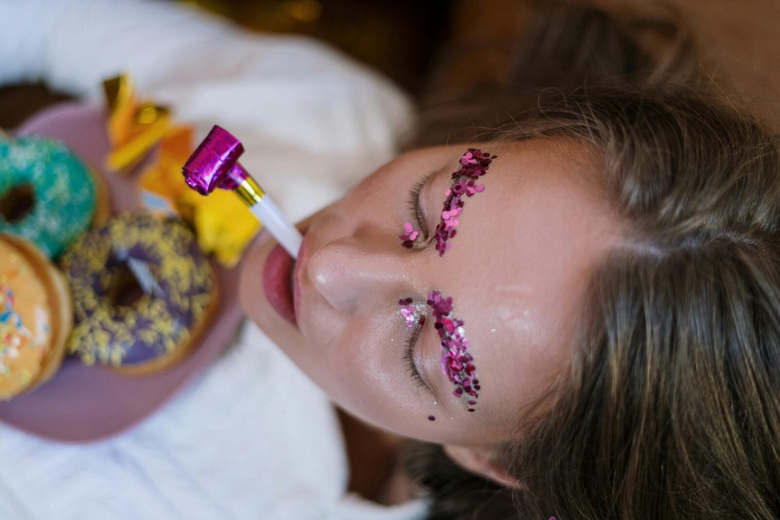 Vibrant makeup and festive scene with glitter eyebrows, party blowers, and tasty doughnuts.